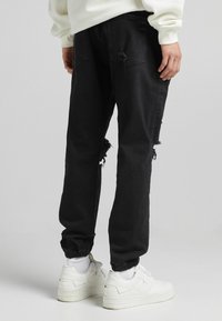 Person som bär svarta slitna jogger-jeans och vita sneakers, sett bakifrån, står på en enfärgad ljusgrå bakgrund.