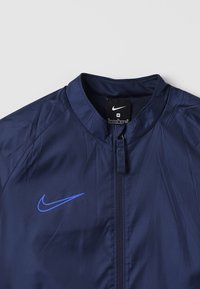 Navy blue windbreaker jacket with a stand collar, front zipper, and a blue Nike swoosh logo. Made from lightweight, smooth material.