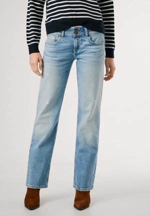 Kobieta nosząca jasnoniebieskie dżinsy o prostych nogawkach z lekkim przetarciem oraz brązowe botki, połączone z czarno-białym swetrem w paski.