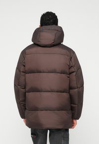 Καφέ πούφφερ μπουφάν με μεγάλη κουκούλα, που διαθέτει ραμμένα τμήματα και λεία υφή. Χωρίς ορατό εξοπλισμό ή αξιοσημείωτες λεπτομέρειες.