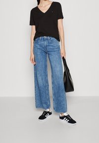 Sort v-hals t-shirt, blå livlig jeans, sorte sneakers med hvide striber og en sort skuldertaske, stående mod en ensfarvet baggrund.