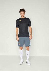 Camiseta atlética negra de manga corta, con un logo dorado en el pecho, combinada con pantalones cortos grises que tienen un emblema rojo. Calcetas altas blancas y zapatos blancos.