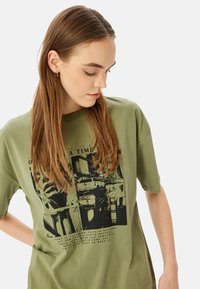 Camiseta de algodón verde oliva con mangas cortas, que presenta una impresión gráfica negra que muestra escenas de la ciudad y texto en la parte delantera.