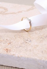 Gouden ring met een centrale heldere steen geflankeerd door kleinere stenen, geplaatst op een gestructureerde lichte stenen ondergrond en een witte strik.