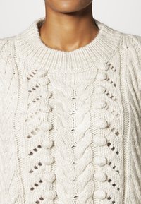 Kremowy sweter dziany z teksturowanym wzorem warkocza, okrągłym dekoltem i ozdobnymi pomponami po bokach. Miękki, gruby materiał.