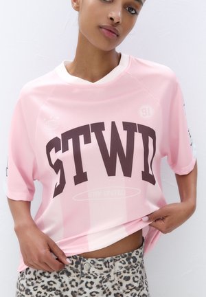 Pink mesh jersey with "STWD" in dark brown on the front. Features short sleeves and a round neckline. Worn over leopard print pants.