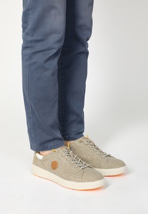 Beige Sneaker mit einem strukturierten Stoffoberteil, hellfarbiger Sohle, gelben Akzenten und einem runden Logo an der Seite, getragen mit marineblauen Hosen.