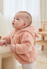 Veste rose douce et duveteuse à capuche avec un tissu texturé. Comprend une fermeture éclair à l'avant et de petites oreilles sur la capuche. Le bébé est appuyé sur une table ronde.