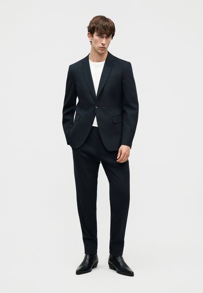 Άνδρας με μαύρο ραμμένο κοστούμι, λευκό t-shirt και μαύρα δερμάτινα μπότες, στέκεται μπροστά σε ένα απλό, ανοιχτό γκρι φόντο.