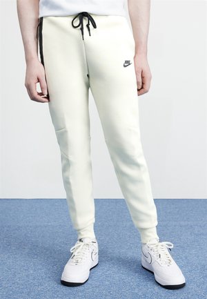 Pantalon de jogging blanc Nike avec cordon noir porté par une personne debout sur un tapis bleu à côté de baskets blanches Nike avec des semelles noires.
