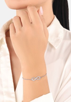 Bracciale d'argento con simbolo dell'infinito, decorato con pietre trasparenti, su un polso, abbinato a una camicia bianca.