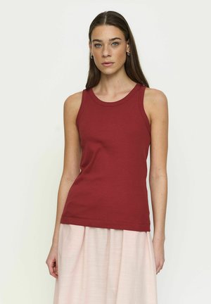 Femme aux longs cheveux bruns portant un haut rouge sans manches et une jupe rose clair, regardant droit devant elle sur un fond uni.