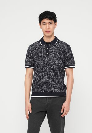 Νεαρός άνδρας που φορά μαύρο και λευκό πόλο με σχέδια και κοντά μανίκια, με μαύρο τζιν, στέκεται μπροστά σε απλό λευκό φόντο.