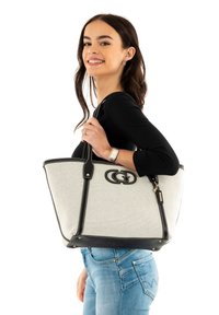 Sac fourre-tout en toile beige avec des garnitures noires, présentant un logo proéminent et deux bretelles solides. Détail de zip en haut.