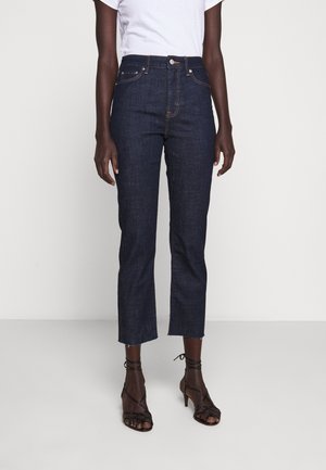 Personne portant un jean court taille haute bleu foncé avec des ourlets bruts et des sandales noires à talons hauts avec brides, sur un fond clair uni.