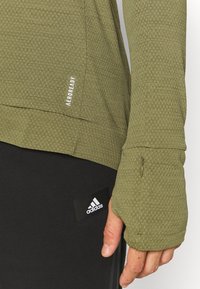 Oliivivihreä urheilullinen pitkähihainen paita, jossa on teksturoitu kuvio, peukalokuoret ja "AEROREADY" -näyttö. Yhdistetty mustiin housuihin, joissa on Adidas-logo.