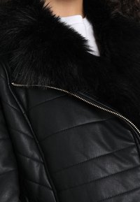 Chaqueta de cuero negra con diseño acolchado, con un collar de piel negra suave y detalle de cremallera plateada. La textura parece lisa y brillante.