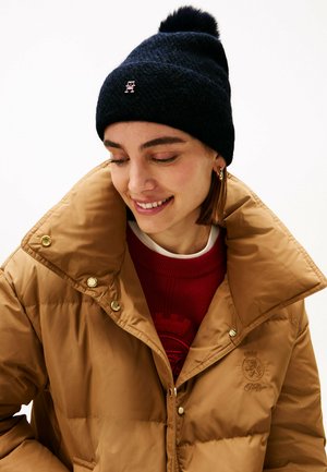 Navy knitted beanie with a pom-pom on top and a small logo, paired with a brown puffer jacket featuring gold-tone buttons and a red sweater.