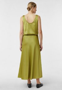 Vrouw met blond haar in een lage knot, gekleed in een mouwloze olijfgroene satijnen jurk en zwarte platte schoenen, staande met haar rug naar de camera op een witte achtergrond.