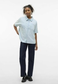 Camicia azzurra chiara a maniche corte con bottoni e due tasche sul petto, abbinata a jeans dritti blu scuro e scarpe nere.
