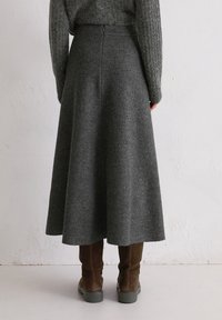 Kobieta ubrana w szaro-włóknianą spódnicę o wysokim stanie, brązowe zamszowe botki do połowy łydki oraz fakturowany szary sweter, stojąca przy białej ścianie.