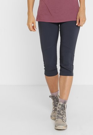 Persoon die marineblauwe capri legging draagt, stoffige roze enkelsokken, beige wandelschoenen en een mauve top, staand op een lichtgrijze vloer.