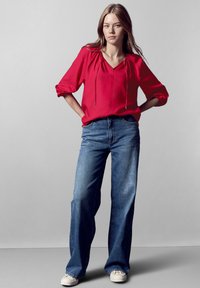 Blusa rossa con dettaglio a cordoncino e maniche 3/4, abbinata a jeans larghi azzurri e sneakers bianche. Texture morbide e vestibilità casual.