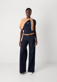 Marine t-skjorte med broderte detaljer, beige strikkegenser drapert over skuldrene, mørk denim bukser med vide ben og hvite sko.