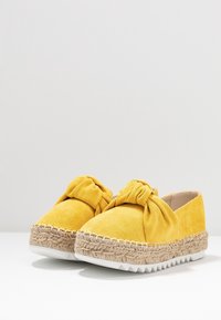 Zapatos amarillos de ante sin cordones con un gran lazo anudado en la parte superior y suelas de plataforma tejidas de yute con bottoms de goma blanca.