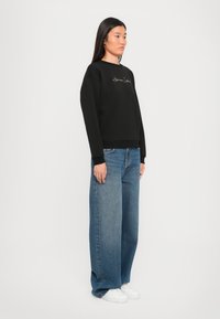 Zwart sweatshirt met gouden geborduurde tekst, gecombineerd met wijde blauwe jeans en witte sneakers. Model staat tegen een neutrale achtergrond.
