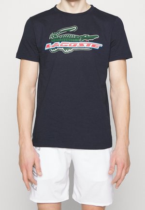 Hombre con una camiseta Lacoste azul marino con un gran logo de cocodrilo verde y pantalones cortos blancos con letras laterales.