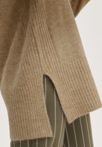 Pull en tricot beige avec texture côtelée, présentant des fentes latérales. Associé à un tissu à rayures vertes et blanches. Apparence douce et chaleureuse.