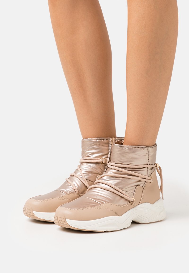 Bas des jambes portant des bottines rembourrées beiges et brillantes avec des semelles blanches épaisses et des lacets enroulés sur un fond uni.