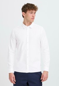 Camisa blanca de manga larga con cuello clásico, hecha de algodón. Presenta un diseño elegante y un dobladillo recto, combinada con pantalones azul marino.