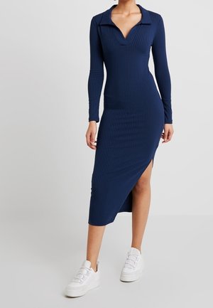 Kvinde iført en marineblå ribstrikket midi-kjole med lange ærmer, krave, V-hals, sideslidser og hvide sneakers, stående mod en ensfarvet baggrund.