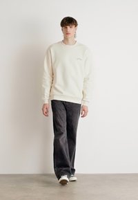 Sudadera crema con puños acanalados, combinada con jeans de pierna ancha en gris oscuro y zapatillas blancas, llevada por un modelo de pie contra un fondo neutro.