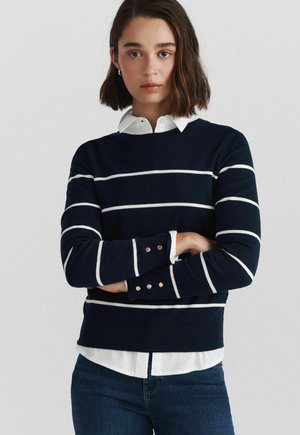 Ung kvinde med skulderlangt mørkt hår iført en navyblå sweater med hvide vandrette striber over en hvid skjorte med krave, armene krydset.