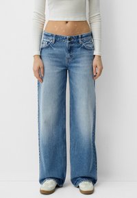Jeans din denim albastru cu picioare largi, cu un finisaj uzat, design cu cinci buzunare și închidere cu buton metalic. Asortați cu adidași albi pentru contrast.