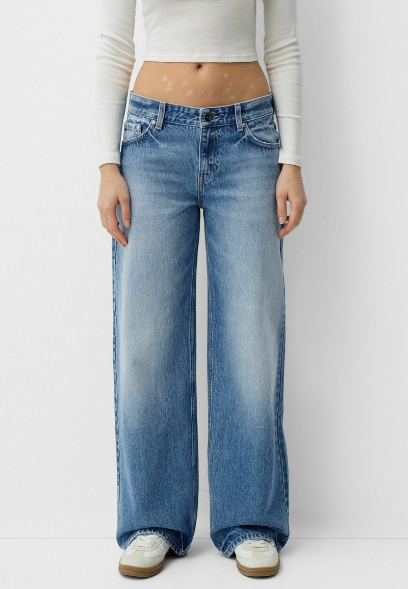 Jeans din denim albastru cu picioare largi, cu un finisaj uzat, design cu cinci buzunare și închidere cu buton metalic. Asortați cu adidași albi pentru contrast.