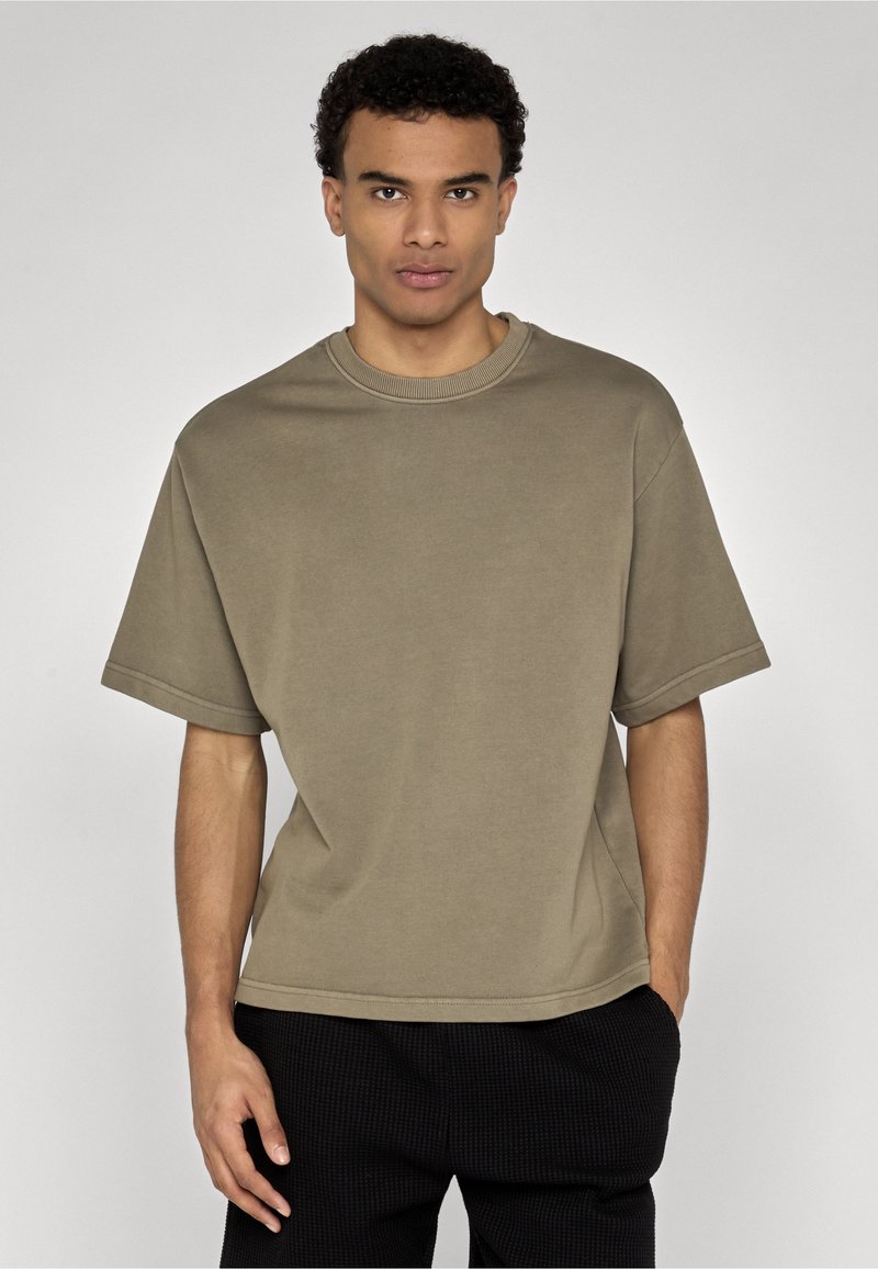 Jeune homme aux cheveux courts et bouclés portant une chemise ample à manches courtes de couleur vert olive et un pantalon noir, se tenant devant un fond uni.