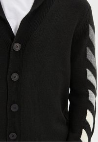 Cardigan nero lavorato a maglia con scollo a V, cinque bottoni neri e una striscia a righe grigia e crema sul braccio destro. Texture morbida.