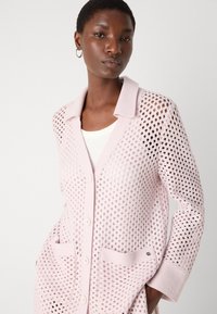 Lichtroze gebreide cardigan met open geweven ontwerp, knoopsluiting en twee voorzakken. Heeft een gekantelde kraag en subtiele logodetails.