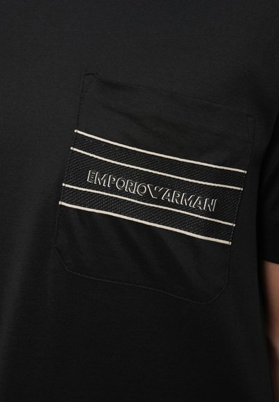 T-shirt preta com um bolso no peito, apresentando riscas horizontais brancas e "EMPORIO ARMANI" impresso em prata numa tecido texturizado.