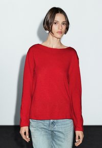 Ein Model mit kurzen Haaren trägt einen leuchtend roten Pullover und helle Jeans und steht vor einem schlichten weißen Hintergrund mit einem dunklen Schatten darunter.