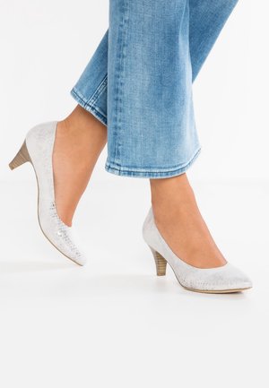 Fødder iført hvide, teksturerede læderpumps med mellemhøj hæl, stylet med lyseblå, korte jeans mod en hvid baggrund.