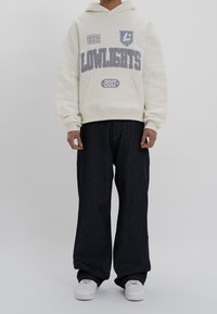 Sweat à capuche crème avec des graphismes gris, incluant "LOWLIGHTS" et un logo ; associé à un jean large foncé et des baskets blanches.