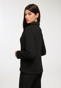 Blazer nero su misura con colletto strutturato, maniche lunghe e tasche laterali. Texture liscia, design aderente e silhouette semplice.