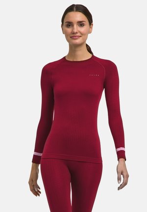 Kvinde iført en tætsiddende langærmet rød træningstop og matchende leggings med pink striber på ærmerne, stående mod en ensfarvet baggrund.