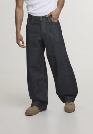 Uomo che indossa jeans a gamba larga blu scuro e scarpe beige con suola spessa, posa con una mano in tasca contro uno sfondo semplice.