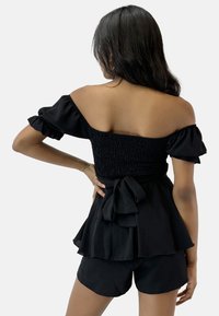Top nero con spalle scoperte, corpino arricciato, maniche corte a volant e fiocco in vita. Abbinato a pantaloni corti neri. Tessuto morbido.